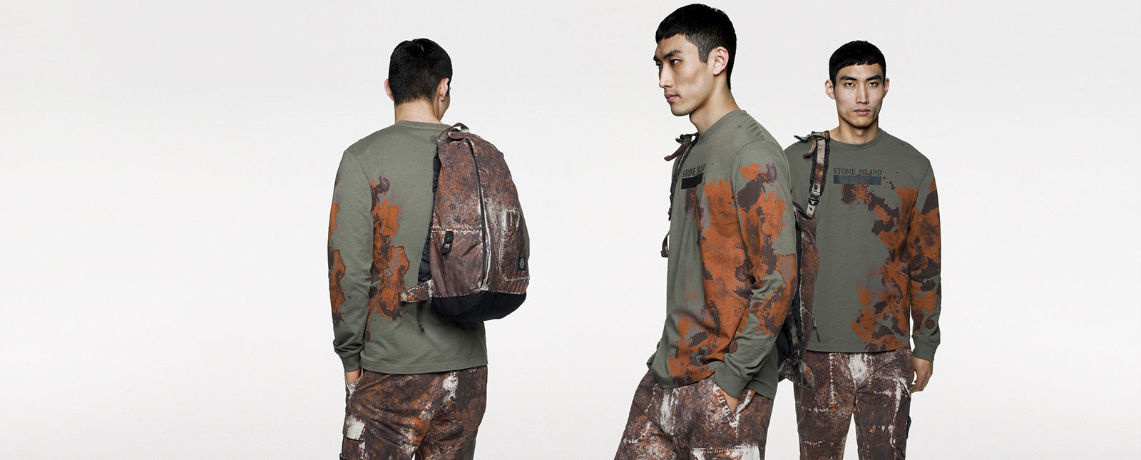 Back, side and front view of model in green, long sleeve T shirt with brown and orange print, carrying a backpack on his shoulder.
