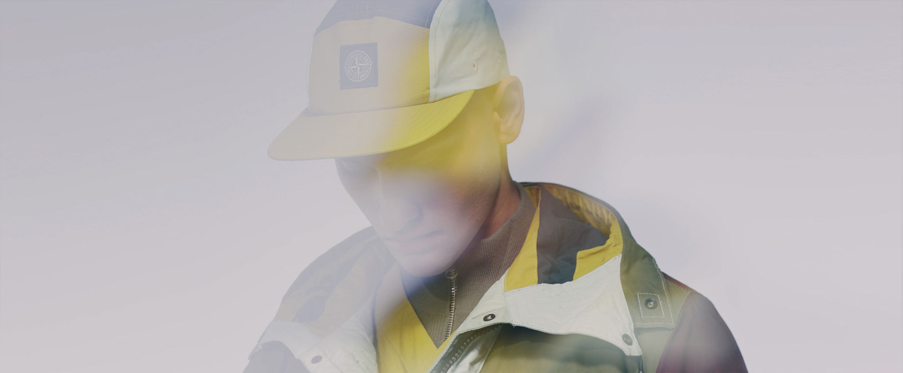 Close up of model wearing multicolored jacket and matching cap wit Stone Island patch.