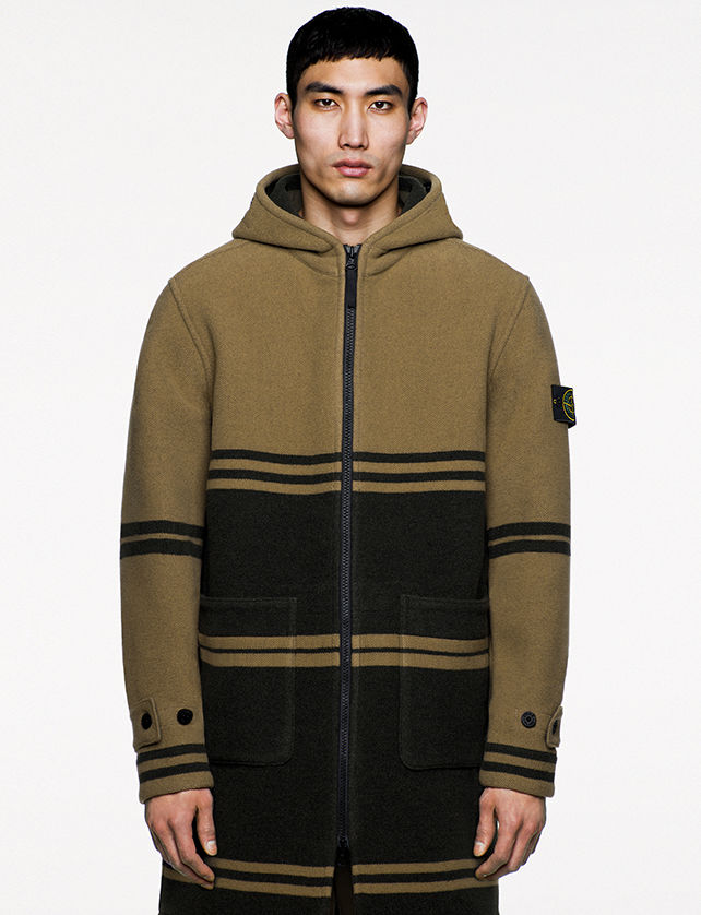 Model wearing olive green and black, thigh length hooded jacket with large horizontal hand pockets and a black zipper fastening.