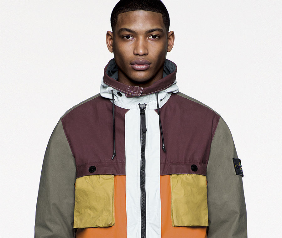 Model wearing multicolored jacket with black zipper fastening and Stone Island badge on left arm.