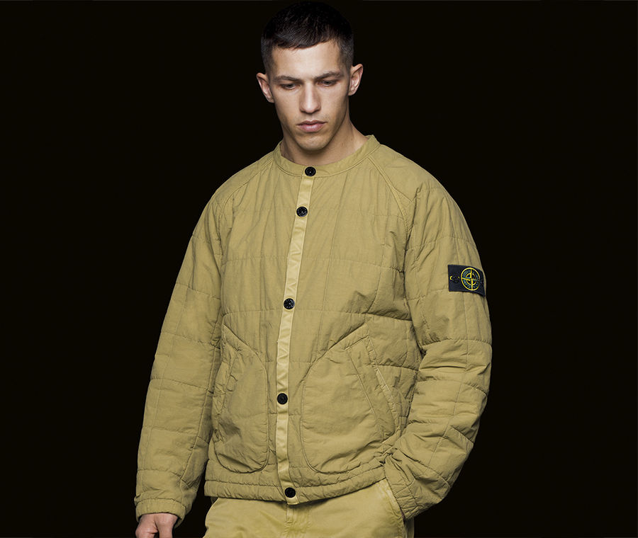 Model wearing jacket in quilted, mustard fabric, with Stone Island badge on left arm.