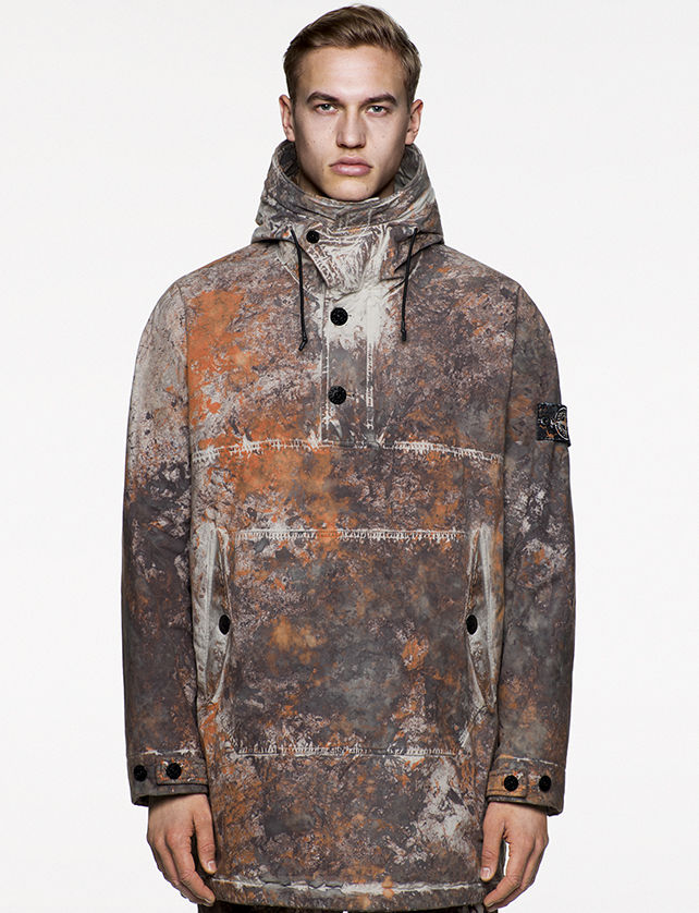 Model wearing multi colored, hooded jacket with button opening at chest and a large pouch pocket.