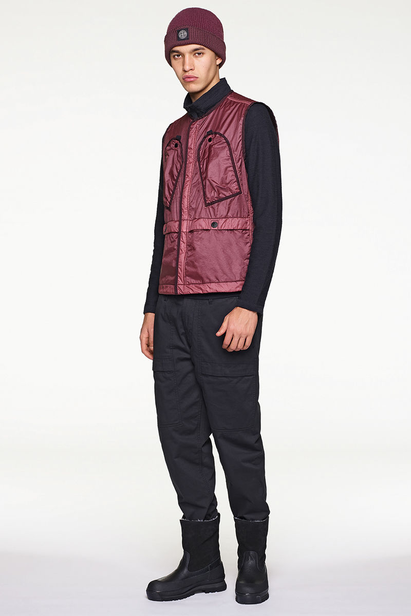 Front view of model in dark burgundy beanie and vest worn with black sweater, pants and calf length boots.
