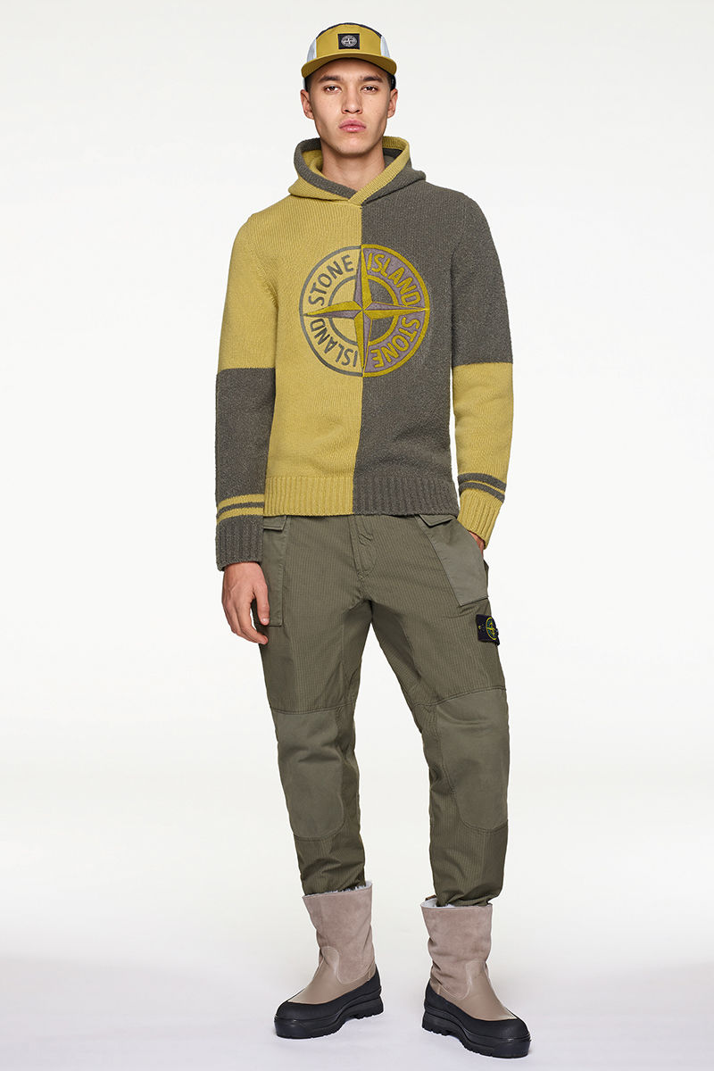 Model wearing mustard and dark green knit sweater with dark green pants, cap and black and sand colored boots.