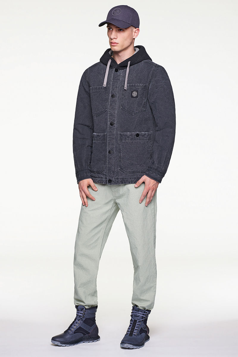 Model wearing dark colored jacket over hoodie, off white pants, cap and black, high top sneakers.