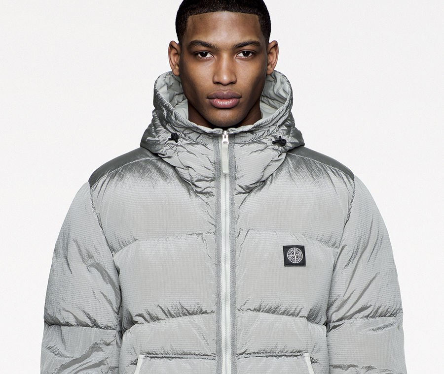 Model with arms folded wearing a silver puffer jacket with high collar and hood and Stone Island patch on the left side of the chest.