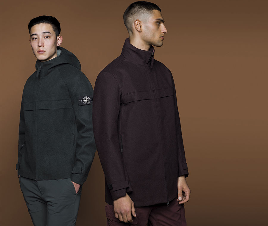 Two models wearing different styles of jackets in dark colors, one with hood and the Stone Island badge on upper left arm and the other with high collar.