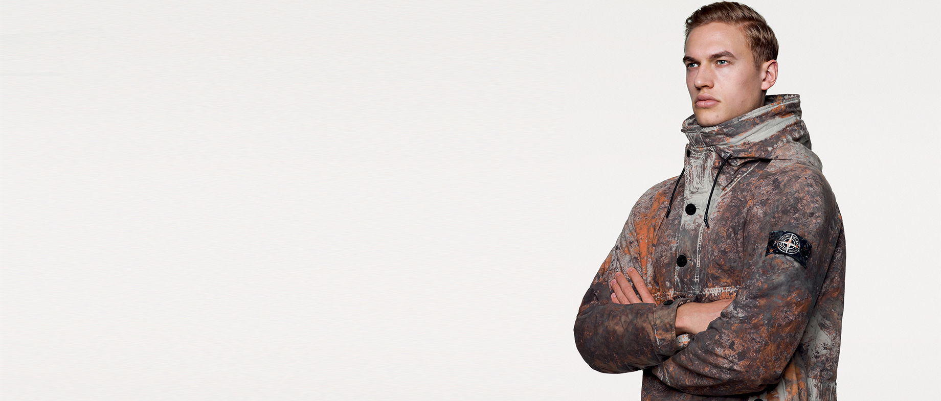 Portrait shot of model wearing hooded technical jacket with multicolored camouflage spatters in earth tones, black buttons on the front and the Stone Island badge on upper left sleeve.