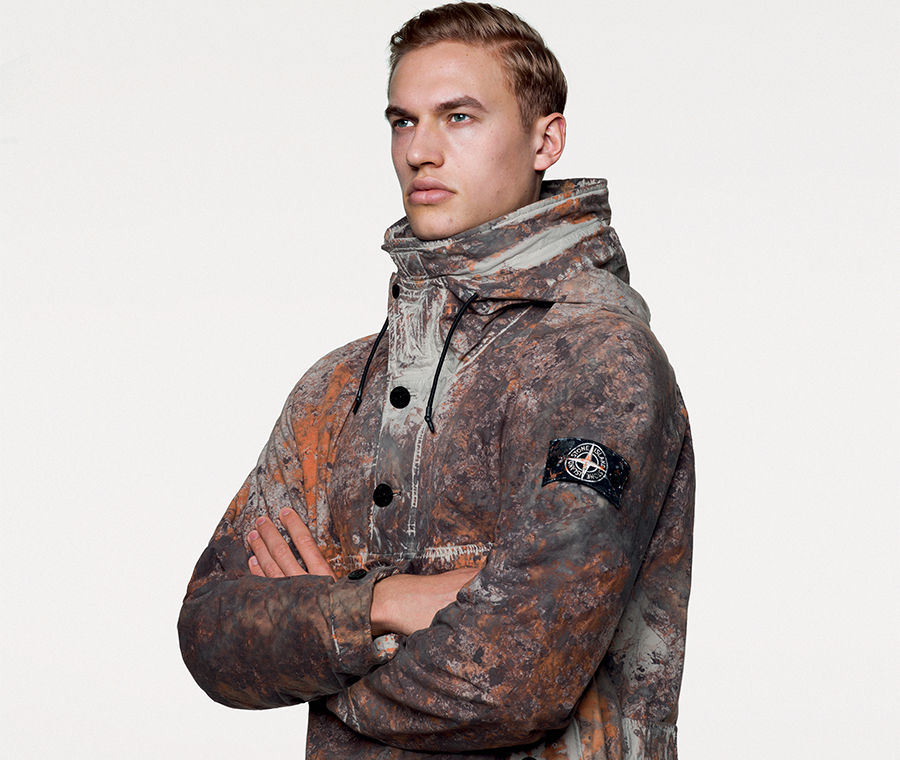 Portrait shot of model wearing hooded technical jacket with multicolored camouflage spatters in earth tones, black buttons on the front and the Stone Island badge on upper left sleeve.