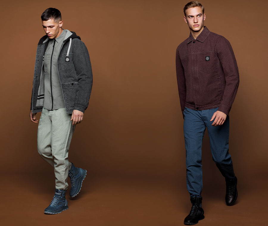 Two models, one wearing a dark grey hooded parka and ankle boots, grey shirt and pants, the other wearing a burgundy collared jacket, blue pants, and black ankle boots