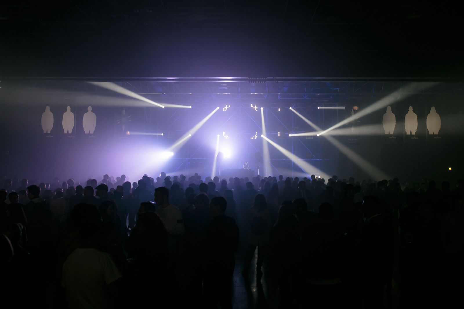Crowd watching a stage with various spotlights shining down and banners with white silhouettes above it
