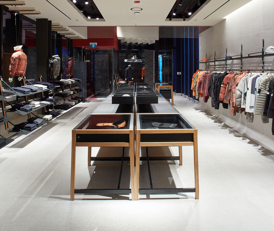 Interior view of the store facing the front entrance with jackets arranged by color hanging on a rack on the right, a display table in the center, folded clothes and mannequins facing windows on the left