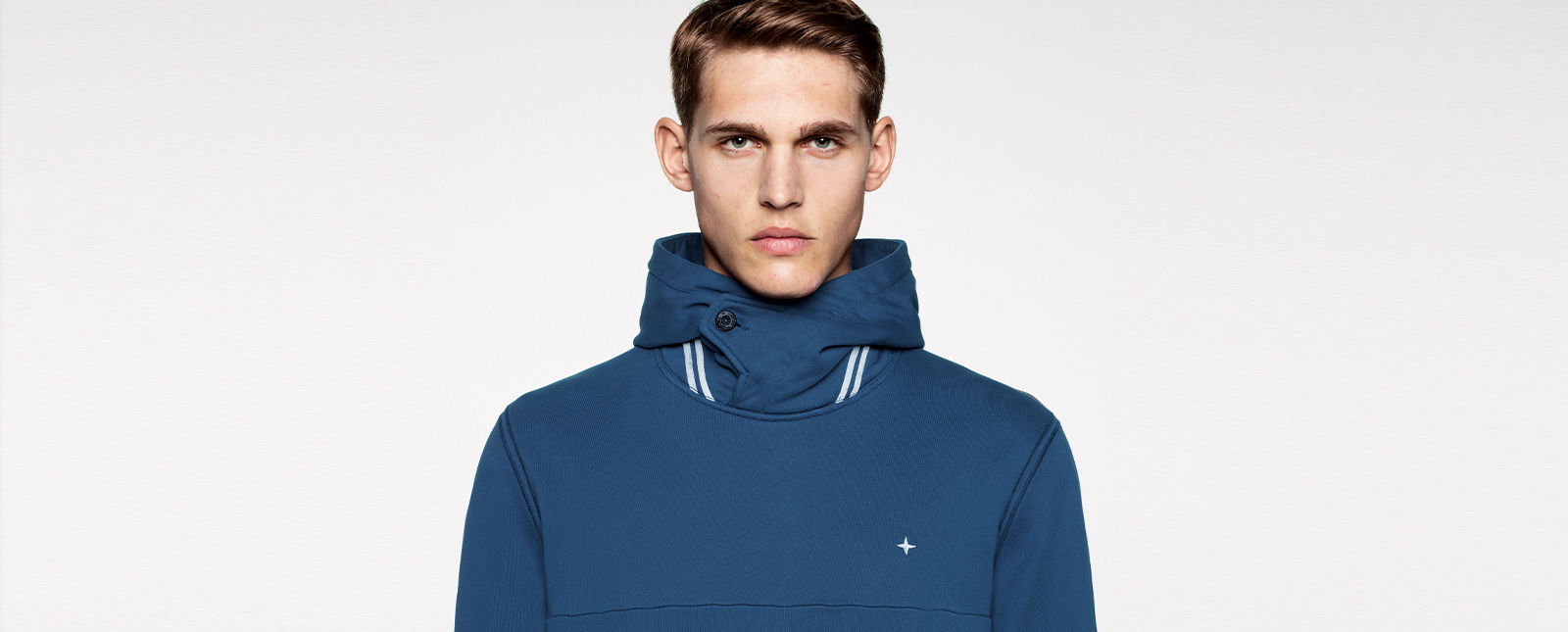 Portrait shot of a model wearing a blue jacket with white stripe details on the buttoned stand collar, with the Stone Island stella on the chest.