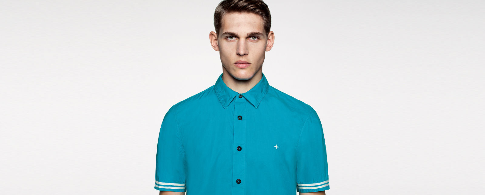 Portrait shot of a model wearing a blue short sleeved shirt with black buttons, white stripe details on the sleeves, and a white Stone Island stella on the chest.