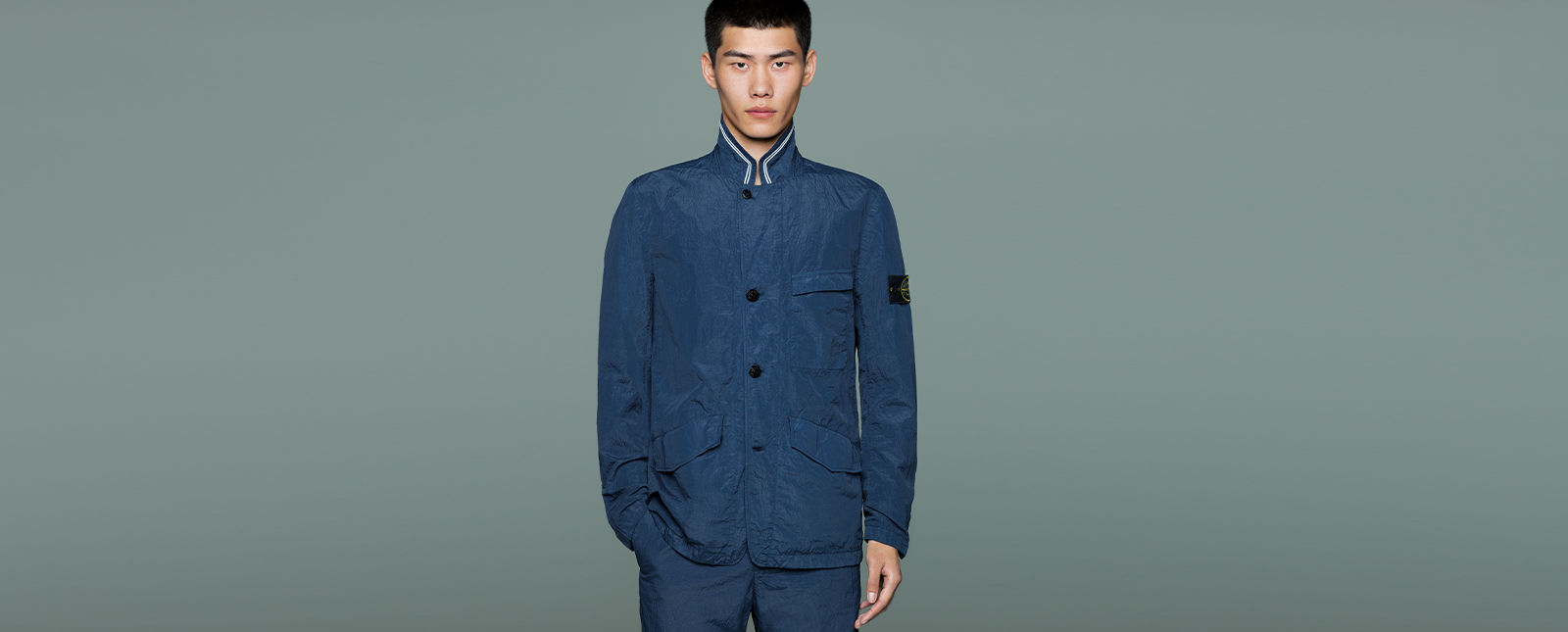 Model wearing dark blue jacket with Mandarin collar with white striped trim, black buttons down the front, chest pocket, side pockets, together with matching pants.