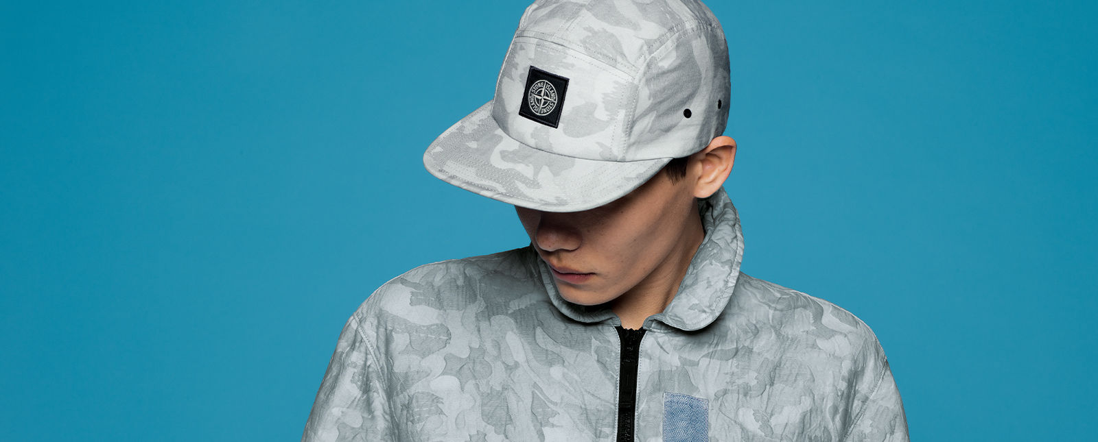 Model wearing light gray camouflage print baseball cap with the Stone Island patch on the front and matching jacket with black zipper closure.