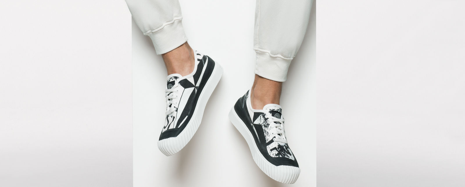 Black and white abstract printed sneakers with thick white rubber sole and white laces on the feet of a model wearing white sweatpants.