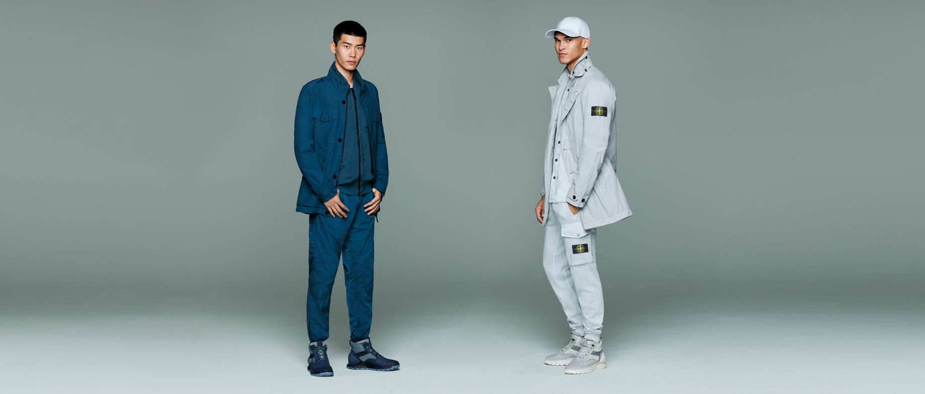 Two models in monochrome outfits, one wears a blue button up jacket over a matching zip up shirt and pants, the other wears a light gray cap, jacket with Stone Island badge on upper left sleeve, button down shirt, and cargo pants with Stone Island badge