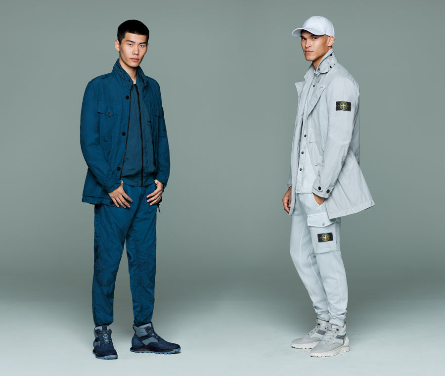 Two models in monochrome outfits, one wears a blue button up jacket over a matching zip up shirt and pants, the other wears a light gray cap, jacket with Stone Island badge on upper left sleeve, button down shirt, and cargo pants with Stone Island badge