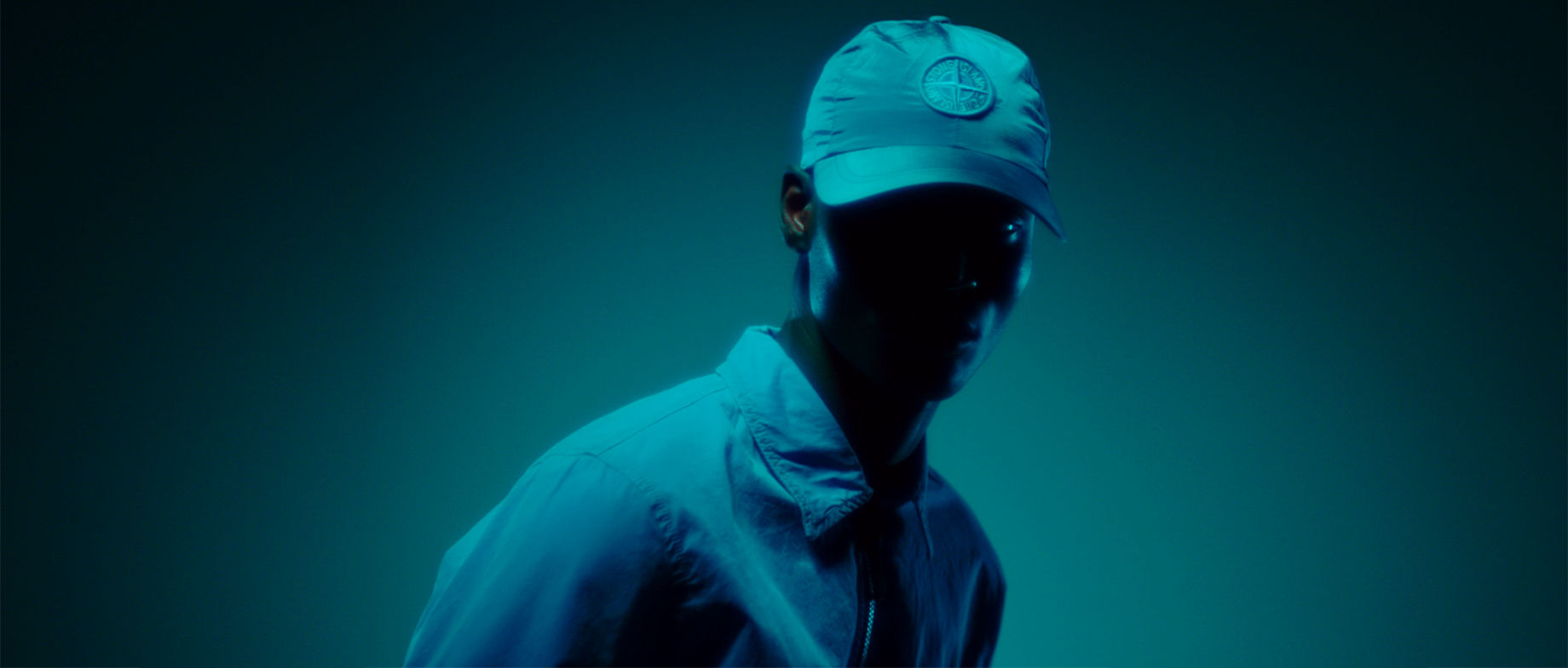 Model standing in shadow wearing a collared shirt jacket and a cap with the Stone Island compass rose.
