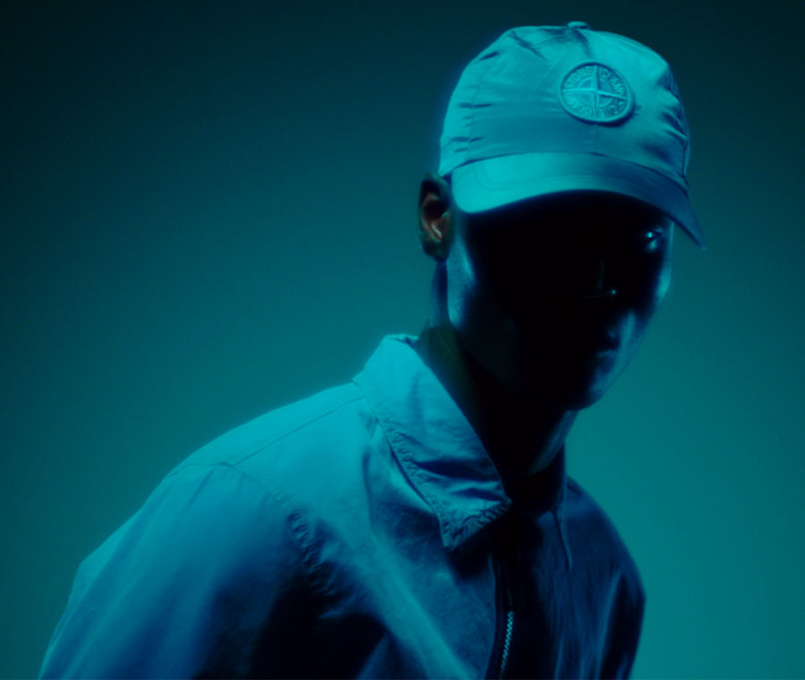 Model standing in shadow wearing a collared shirt jacket and a cap with the Stone Island compass rose.