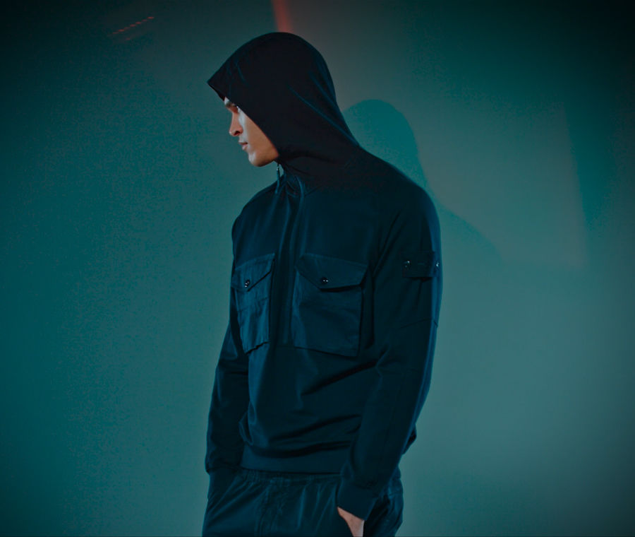Model wearing dark blue jacket with high collar, hood, flap pockets with button closure on the chest and Ghost Badge on the upper left arm.