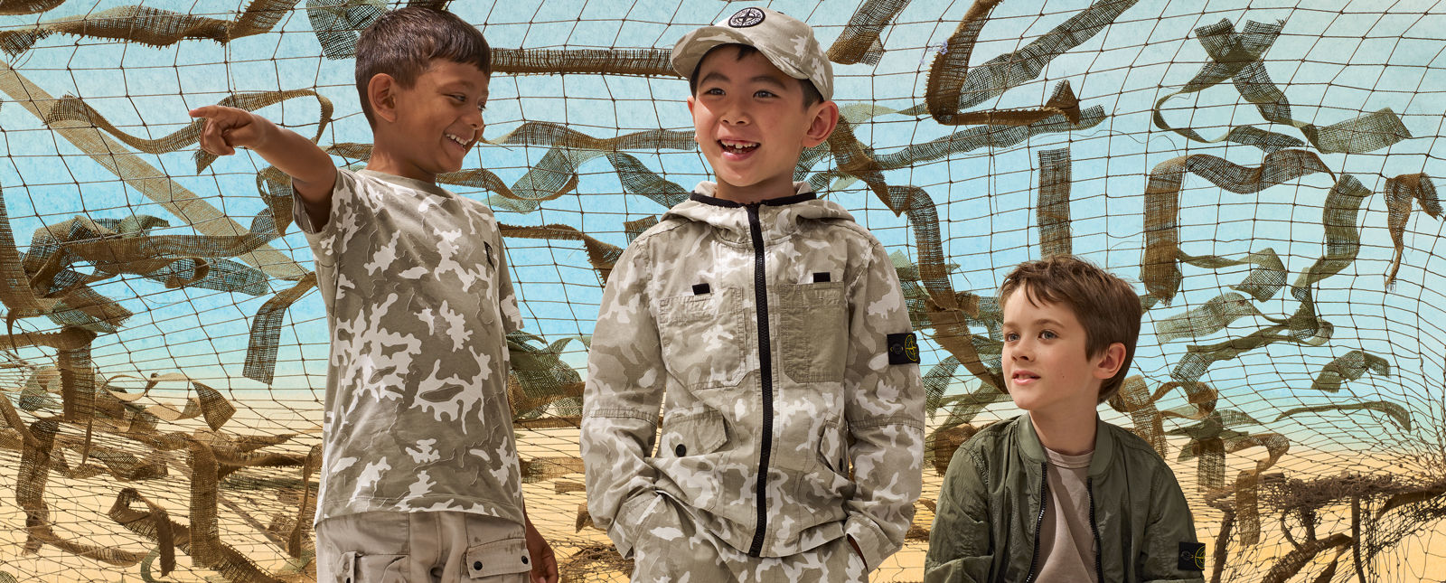 Three junior models, one wearing khaki cargo pants and a camouflage t shirt, another wearing a camouflage jacket with matching pants and cap, and the third wearing a khaki t shirt and military green bomber jacket.