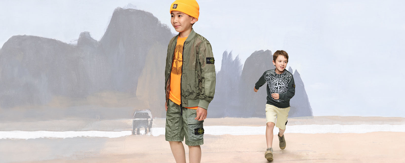Two junior models, one wearing military green cargo shorts and jacket, a yellow top and beanie, the other one wearing cream shorts and a dark colored sweatshirt with the Stone compass rose across the chest.