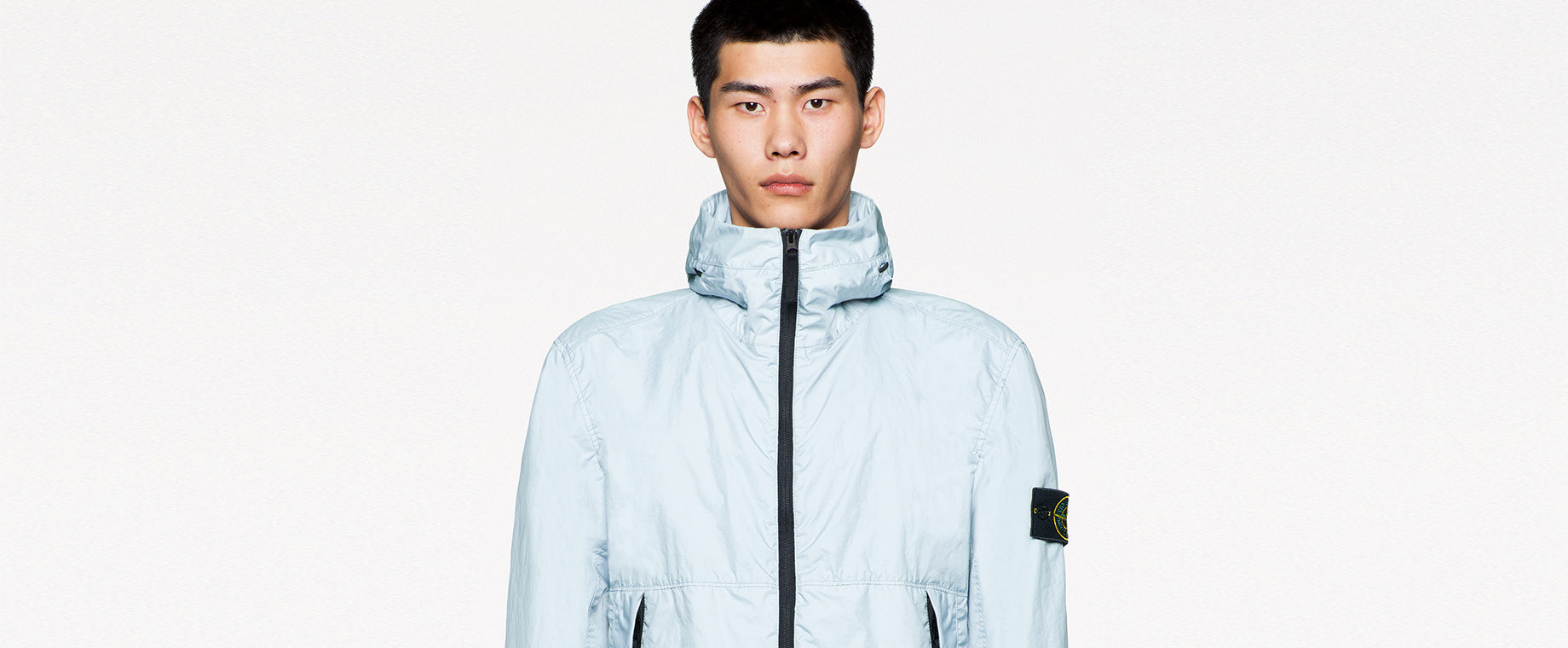 Model wearing dark gray pants and high collared light blue jacket with black zipper down the front and two pockets with zipper closure.