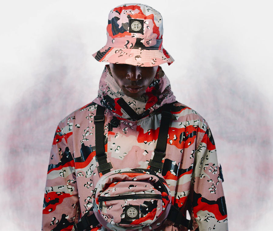 Model wearing a high collar jacket, a bucket hat and a chest pouch with black straps across the shoulders and waist, all in red, pink and off white camouflage print.