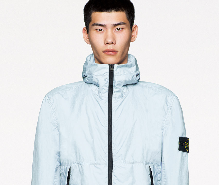 Model wearing dark gray pants and high collared light blue jacket with black zipper down the front and two pockets with zipper closure.