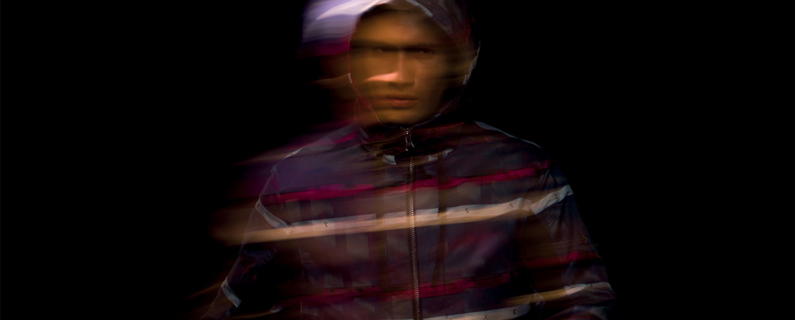 Blurred portrait of model wearing dark colored hooded jacket with white, black and red stripes and zipper closure.