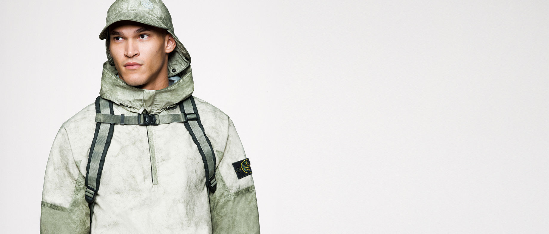Model wearing a parka with high collar, a cap with neck flap and a backpack with sternum strap, all in a matching khaki and green pattern.
