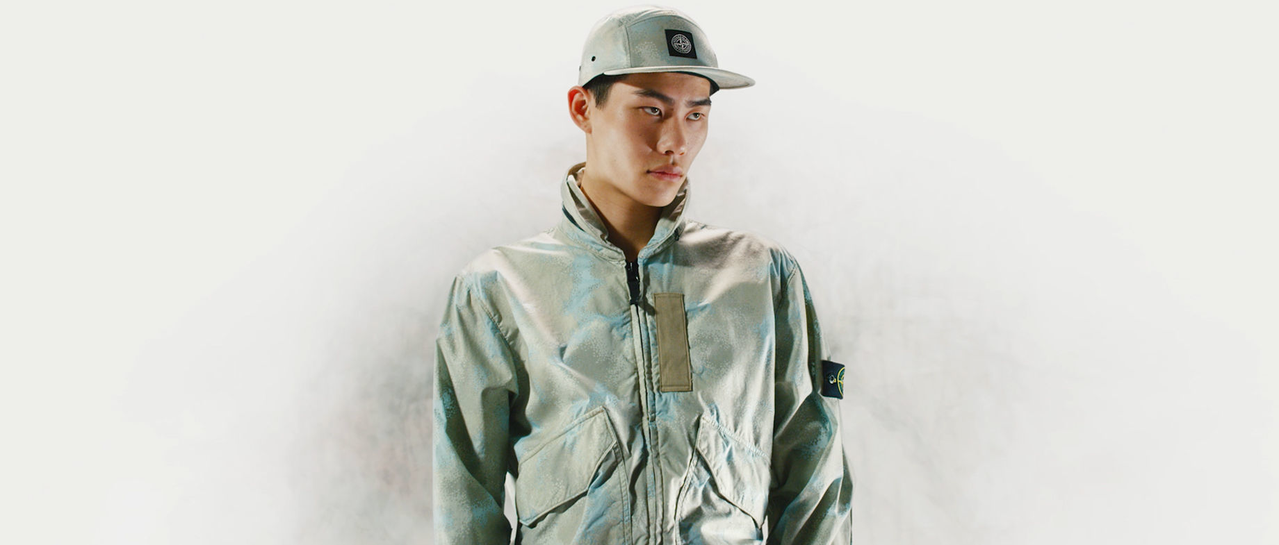 Model wearing a cap and a jacket with high collar, zipper closure and patch pockets, both in the same khaki and light blue camouflage print.
