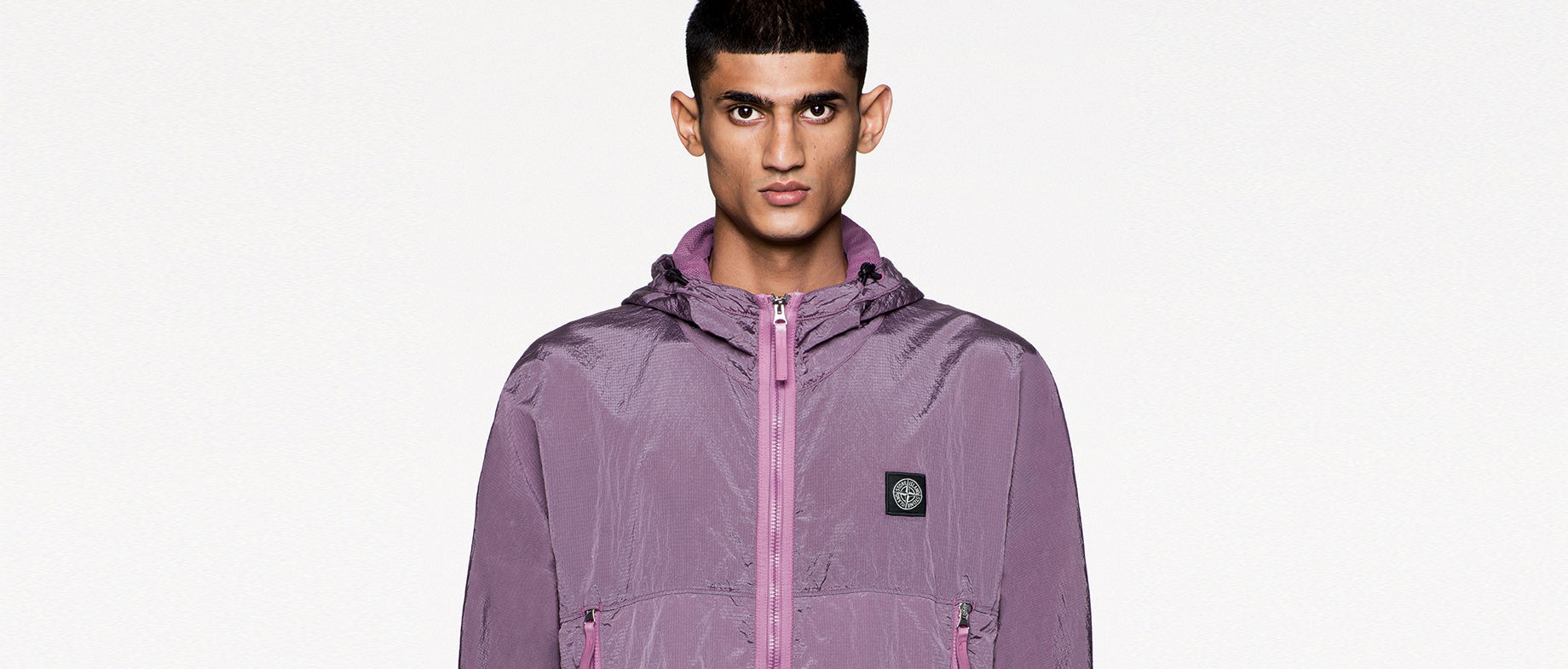 Model wearing a hooded purple jacket with zipper closure, zippered side pockets and Stone Island patch at left chest.