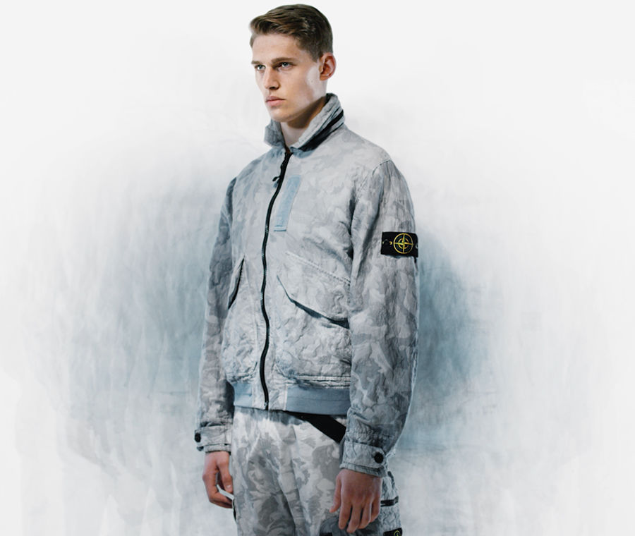Model wearing cargo pants and a bomber jacket with high collar, zipper closure and flap pockets, both in the same gray camouflage print.