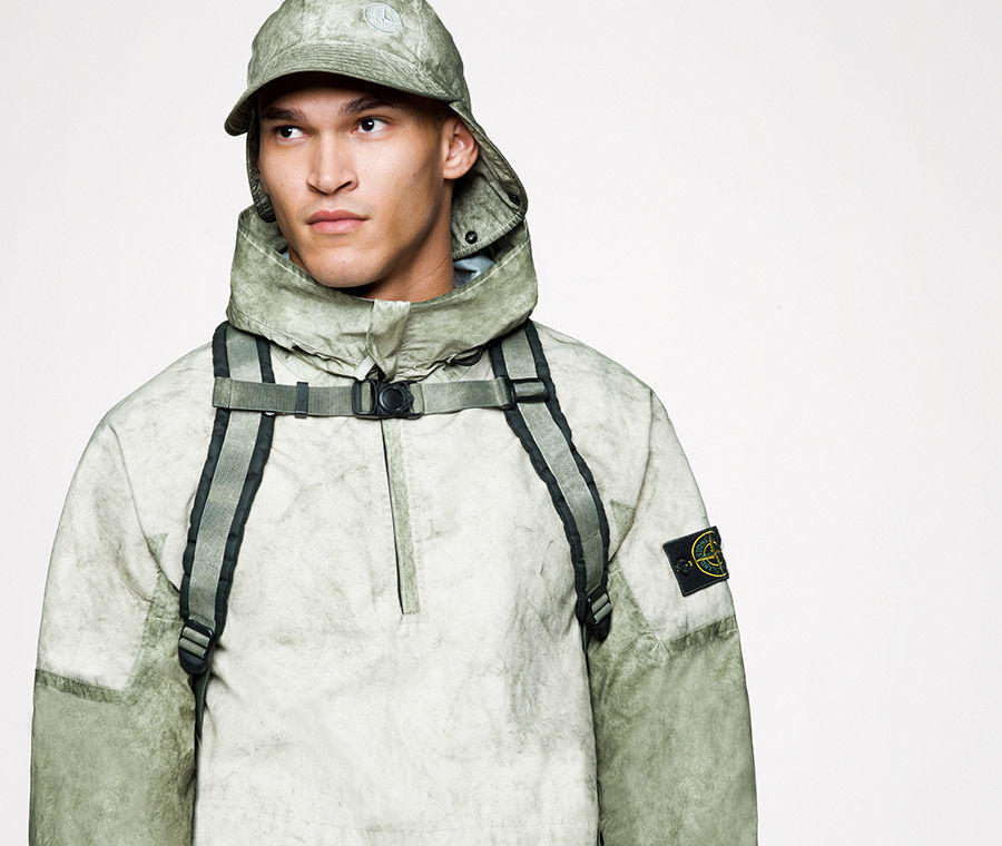 Model wearing a parka with high collar, a cap with neck flap and a backpack with sternum strap, all in a matching khaki and green pattern.