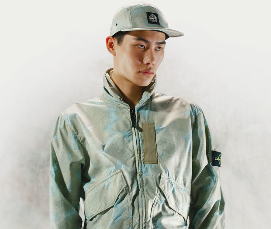 Model wearing a cap and a jacket with high collar, zipper closure and patch pockets, both in the same khaki and light blue camouflage print.