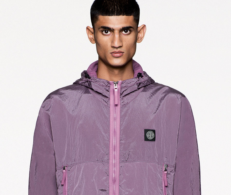 Model wearing a hooded purple jacket with zipper closure, zippered side pockets and Stone Island patch at left chest.