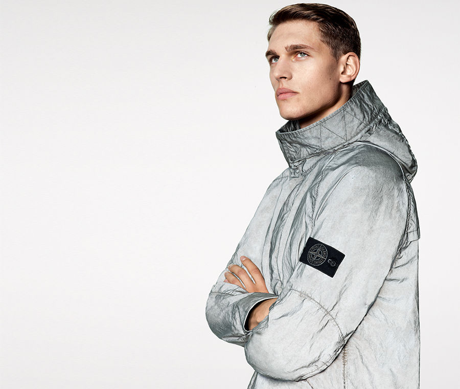 Model wearing a light gray reflective jacket with hood and Stone Island badge on upper left arm.
