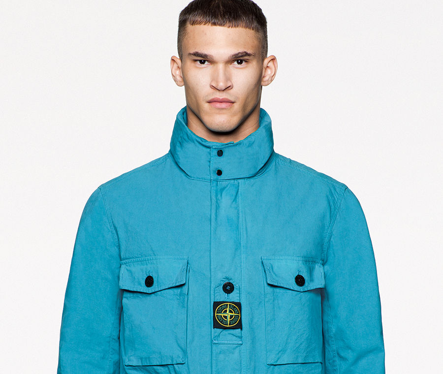 Model wearing a bright blue jacket with stand collar, four flap pockets on the front with black buttons, Stone Island patch in the center of the chest