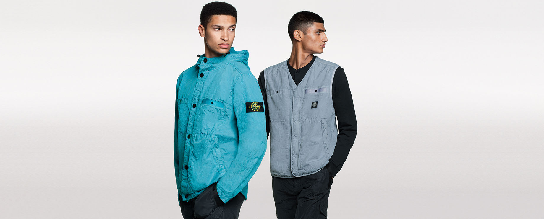 Two models, one wearing black pants and a bright blue hooded jacket with button closure, two patch pockets and two side pockets, the other one wearing black pants, a black shirt and a gray V neck vest with two patch pockets and two side pockets.