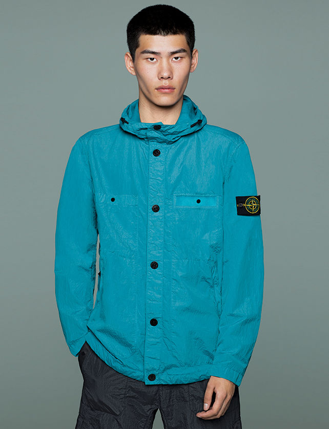 Model wearing black pants and a bright blue hooded jacket with button closure, two patch pockets, two side pockets and Stone Island badge on upper left arm.