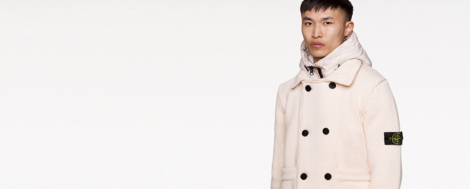 Model wearing an off white knit jacket with collar, hood with zipper, double breasted buttons fastening and the Stone Island badge on upper left arm.