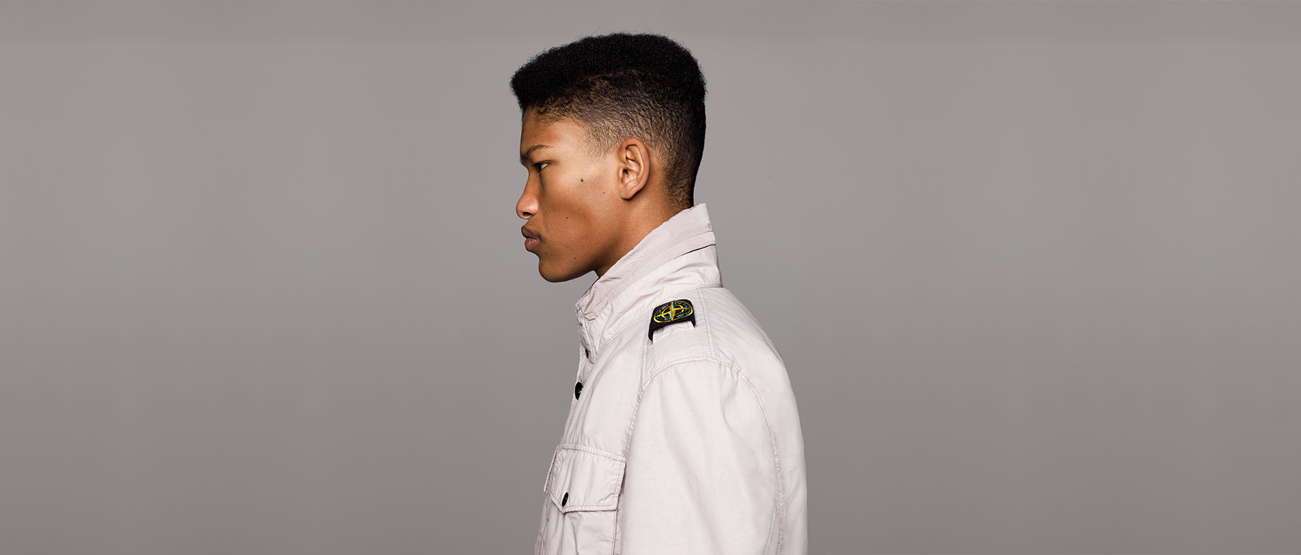 Side view of model wearing an off white jacket with standing collar, bellows chest pocket with flap and snap fastening and Stone Island badge on left shoulder.