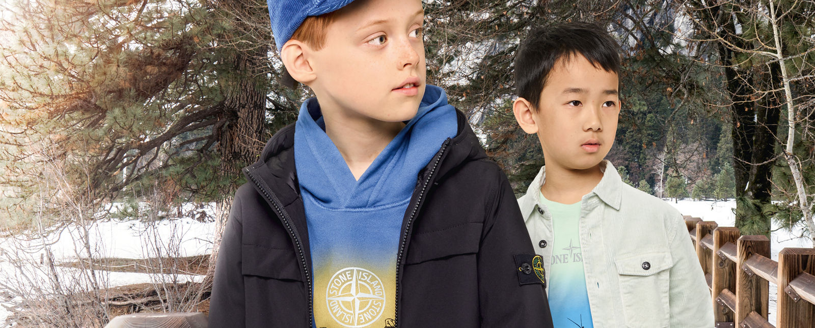Two junior models, one wearing a hooded blue sweatshirt with a colorful print and the Stone Island compass logo, a blue cap and a dark colored jacket with zipper fastening, the other one wearing a light green t shirt with a colorful print on the front and
