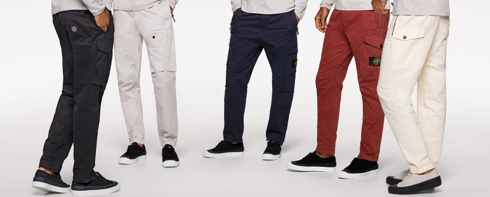 Five models standing in a semi circle and wearing off white tops, sneakers and different styles of cargo pants in black, white, dark blue, brick red and cream.