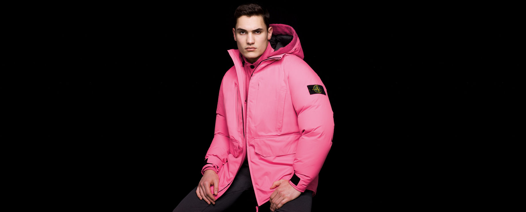 Side view of model wearing black pants and a pink hooded jacket with inner drawstring on back and the Stone Island badge on the upper left sleeve.