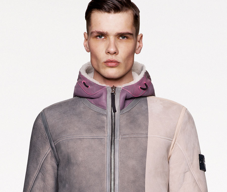 Model wearing dark colored pants and a gray, off white and mauve sheepskin jacket with hood, zipper fastening, two hand pockets and two horizontal black stripes across the left hip area.