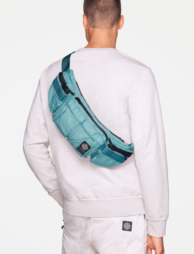 Back view of model wearing a white sweatshirt, white pants with the Stone Island patch on the back pocket and a light blue waist bag with black zippers and multiple pockets, worn over the shoulder.